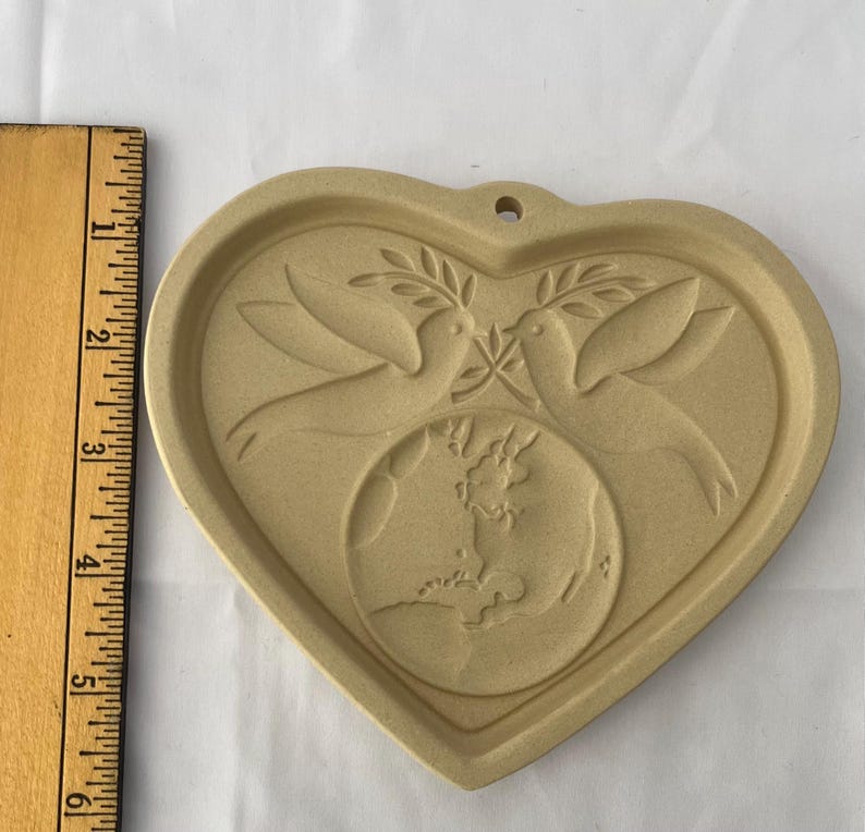 May include: A tan ceramic heart-shaped mould with a design of two doves holding olive branches above a globe. The mould has a small hole at the top for hanging. A wooden ruler is on the left.