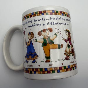 May include: A white ceramic mug featuring a colorful illustration of three children and the text "Touching hearts... inspiring souls... making a difference..." along with the words "FAITH", "JOY", and "HOPE" printed above a checkered border.