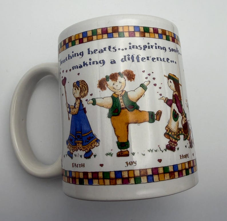 May include: A white ceramic mug featuring a colorful illustration of three children and the text "Touching hearts... inspiring souls... making a difference..." along with the words "FAITH", "JOY", and "HOPE" printed above a checkered border.