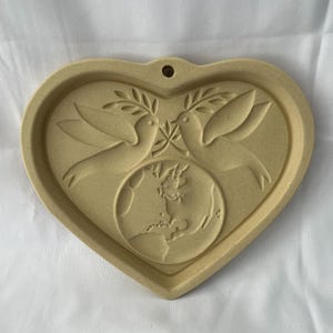 May include: A heart-shaped ceramic wall hanging in a tan colour. The design features two doves holding olive branches above a globe. The heart has a small hole at the top for hanging. The doves and globe are raised.