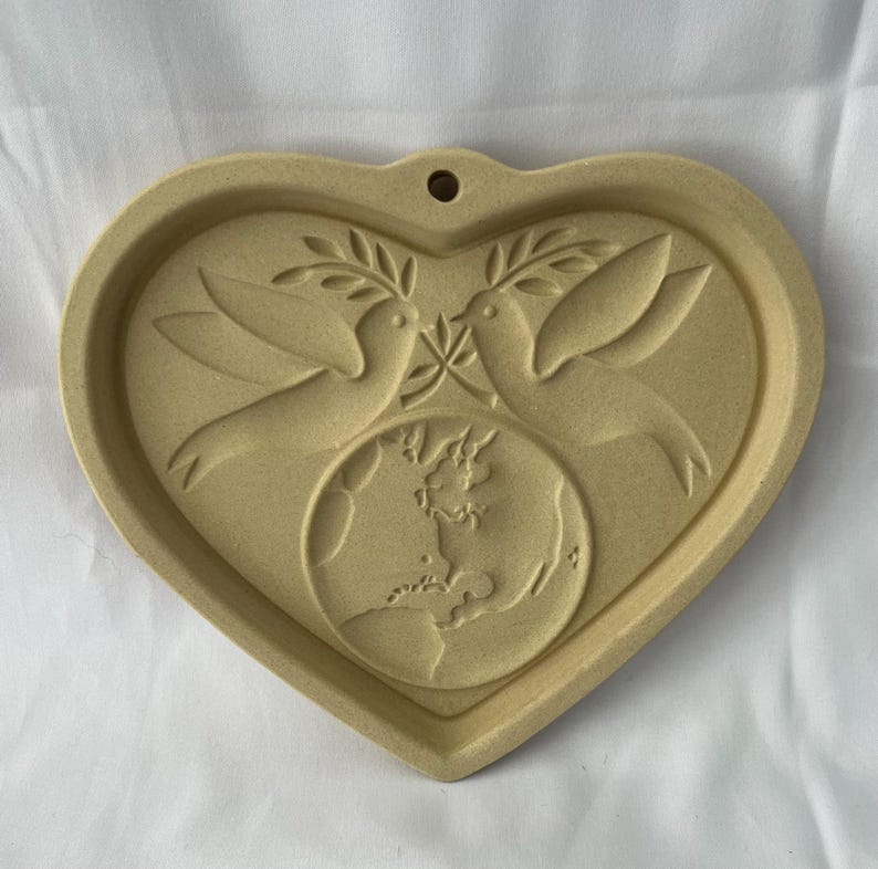 May include: A heart-shaped ceramic wall hanging in a tan colour. The design features two doves holding olive branches above a globe. The heart has a small hole at the top for hanging. The doves and globe are raised.