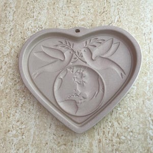 May include: A heart-shaped, light beige decorative plaque. Two doves with olive branches are above a globe. The plaque has a hole at the top for hanging. The design is embossed on the surface.