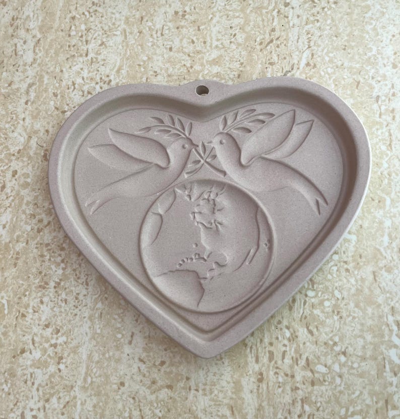 May include: A heart-shaped, light beige decorative plaque. Two doves with olive branches are above a globe. The plaque has a hole at the top for hanging. The design is embossed on the surface.