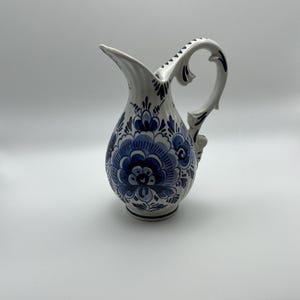 May include: White ceramic pitcher with a blue floral design. The pitcher has a curved handle and a spout. The blue floral pattern covers the body of the pitcher, with additional blue accents on the handle and spout. A decorative piece for the home.