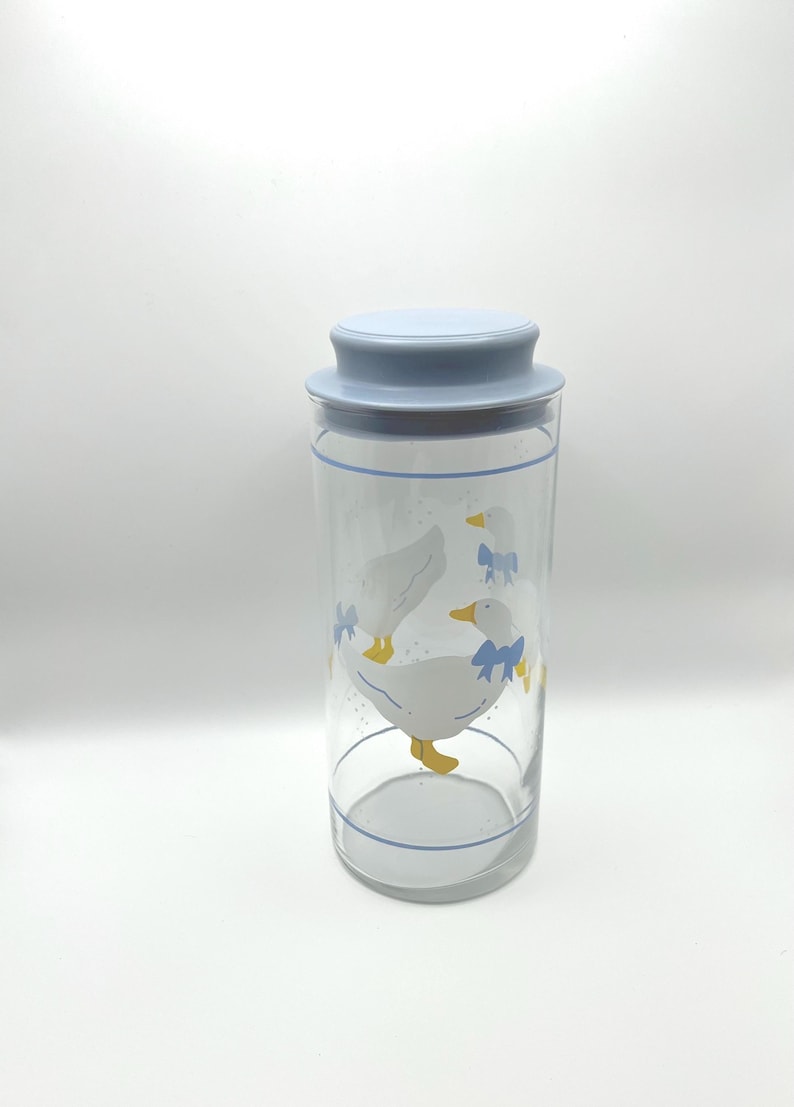 May include: A clear glass jar with a light blue lid and a blue rim. The jar features a design of white geese with yellow beaks and feet, wearing blue bows. The jar is cylindrical in shape.
