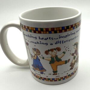 May include: A white ceramic mug featuring a colorful border of small squares and an illustration of three children. The text reads: Touching hearts... inspiring souls... making a difference. Below the children are the words Faith, Joy, and Hope.