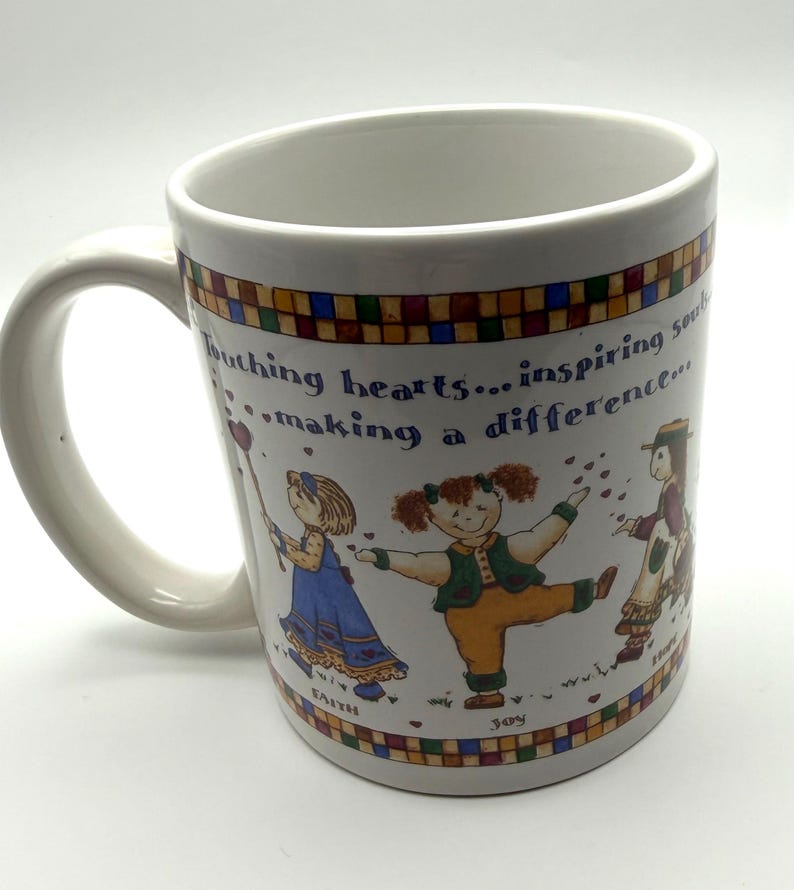 May include: A white ceramic mug featuring a colorful border of small squares and an illustration of three children. The text reads: Touching hearts... inspiring souls... making a difference. Below the children are the words Faith, Joy, and Hope.