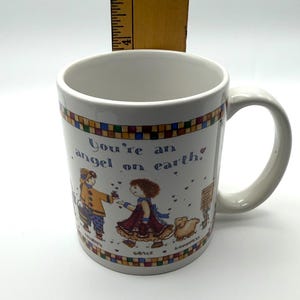 May include: A white ceramic mug featuring a colorful illustration of two figures and a small animal, with the text "You're an angel on earth." A wooden ruler is positioned behind the mug, showing a height measurement of approximately 4.5 inches.