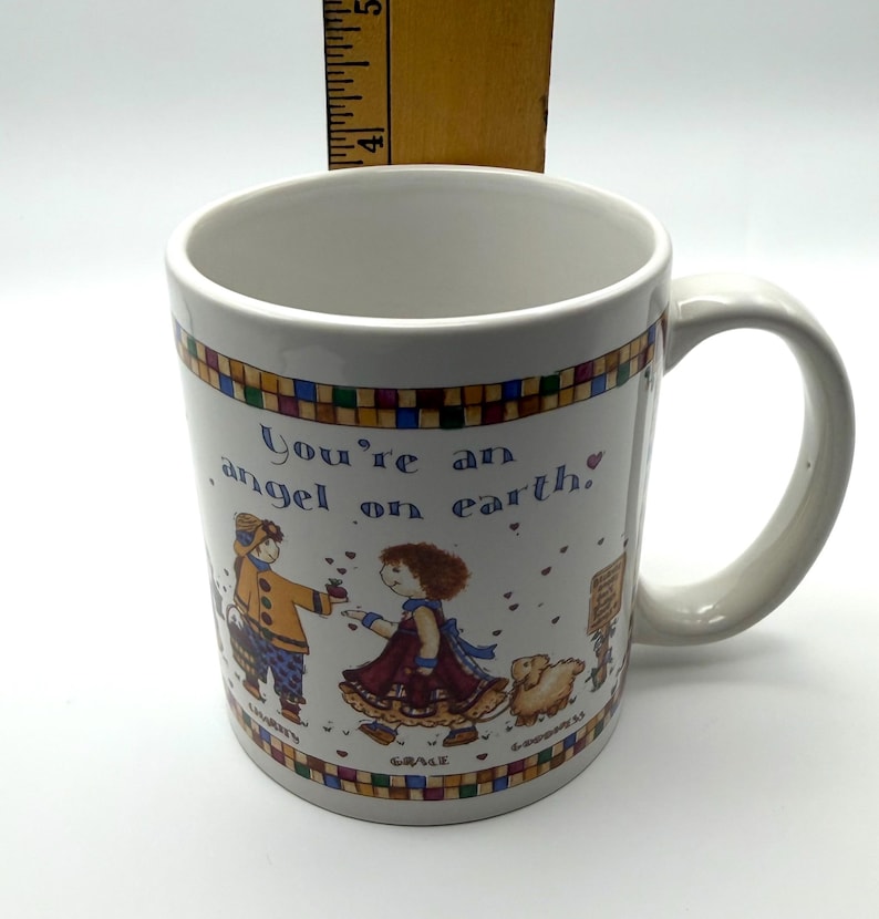 May include: A white ceramic mug featuring a colorful illustration of two figures and a small animal, with the text "You're an angel on earth." A wooden ruler is positioned behind the mug, showing a height measurement of approximately 4.5 inches.