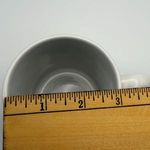 May include: A white ceramic mug viewed from above, positioned behind a wooden ruler that shows the opening of the mug measures approximately 3.5 inches in diameter. The interior of the mug is smooth, white, and circular, with a handle visible on the right.