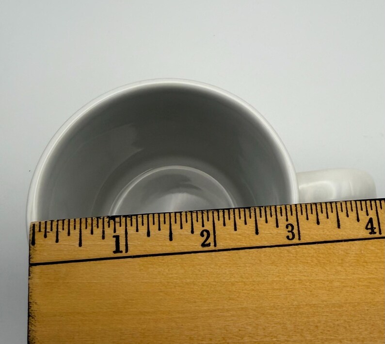 May include: A white ceramic mug viewed from above, positioned behind a wooden ruler that shows the opening of the mug measures approximately 3.5 inches in diameter. The interior of the mug is smooth, white, and circular, with a handle visible on the right.