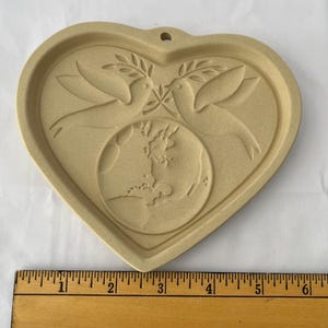 May include: Tan heart-shaped cookie cutter with a dove and olive branch design. Two doves face each other above a globe. The cookie cutter has a small hole at the top for hanging. The ruler next to the cookie cutter measures up to 15 cm.