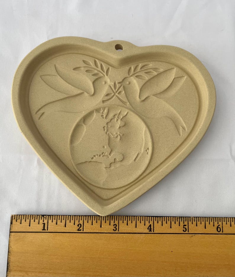 May include: Tan heart-shaped cookie cutter with a dove and olive branch design. Two doves face each other above a globe. The cookie cutter has a small hole at the top for hanging. The ruler next to the cookie cutter measures up to 15 cm.