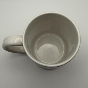 May include: A plain, off-white ceramic mug viewed from directly above, showing the circular opening, the smooth interior walls, and a curved handle extending to the left side. The mug has a simple, cylindrical shape with a glossy finish.