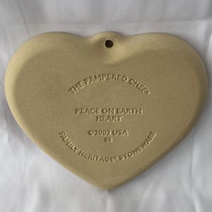 May include: A heart-shaped stoneware piece in a light tan colour. The text "THE PAMPERED CHEF", "PEACE ON EARTH HEART", "&copy;2002 USA SS", and "FAMILY HERITAGE STONEWARE" are inscribed on the surface. A small hole is at the top.