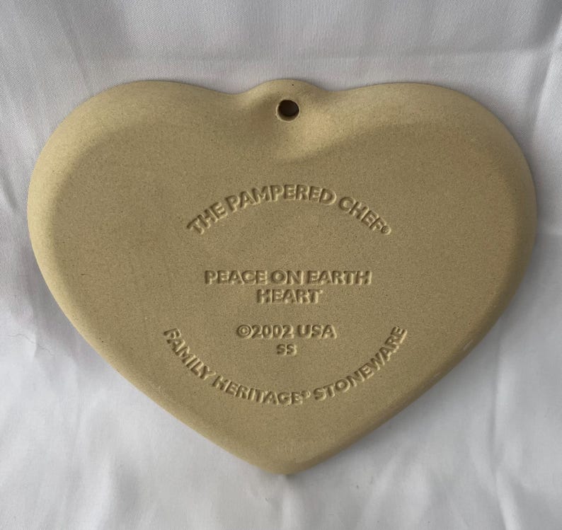May include: A heart-shaped stoneware piece in a light tan colour. The text "THE PAMPERED CHEF", "PEACE ON EARTH HEART", "&copy;2002 USA SS", and "FAMILY HERITAGE STONEWARE" are inscribed on the surface. A small hole is at the top.