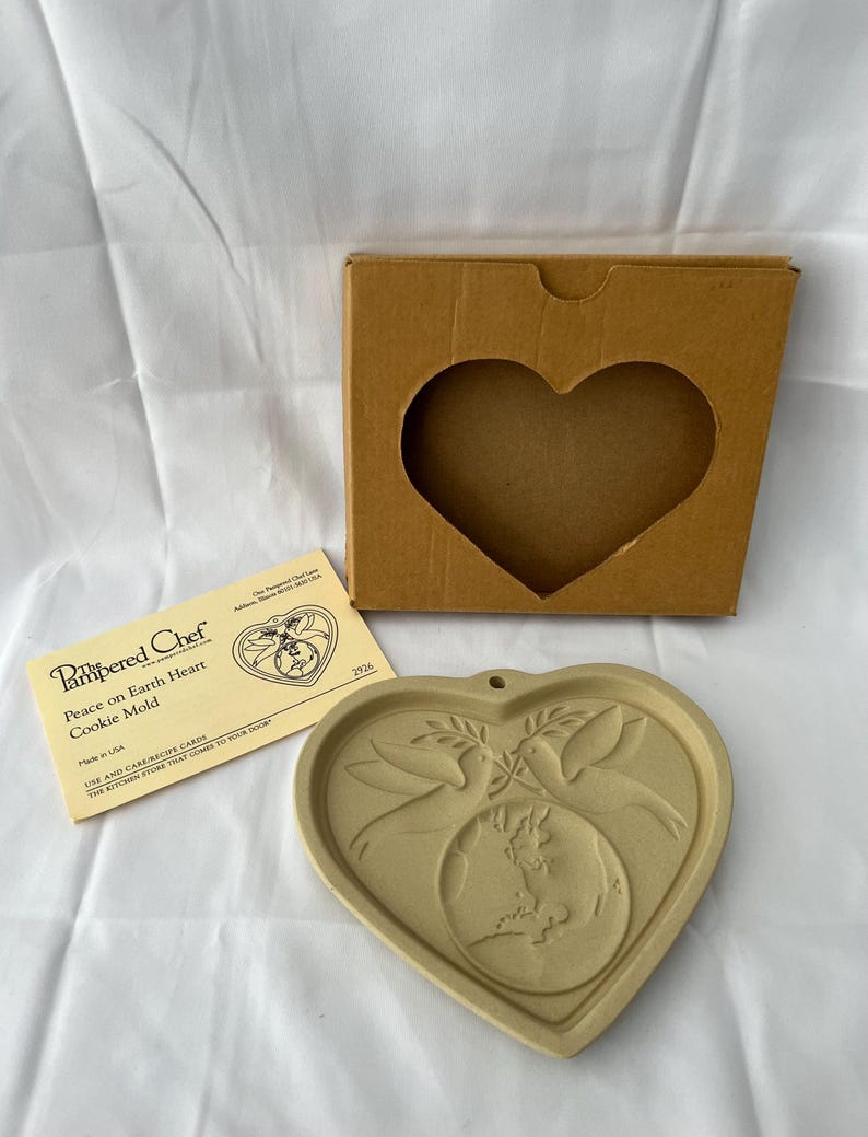 May include: A heart-shaped cookie mold with a dove and globe design, along with its original box and instructions. The mold is beige and features the words "Peace on Earth Heart Cookie Mold".