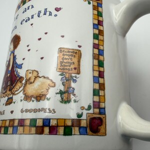 May include: A white ceramic mug featuring a colorful illustration of a child with a lamb, surrounded by a checkered border. The text reads: "Because angels don't always have wings" and "GOODNESS". Small red hearts are scattered across the design.