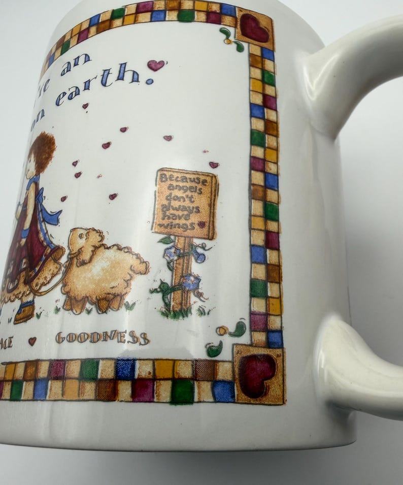 May include: A white ceramic mug featuring a colorful illustration of a child with a lamb, surrounded by a checkered border. The text reads: "Because angels don't always have wings" and "GOODNESS". Small red hearts are scattered across the design.