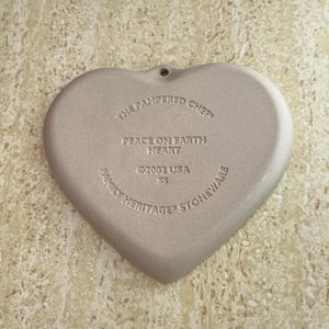 May include: A heart-shaped stoneware piece with the text "The Pampered Chef", "Peace on Earth Heart", and "Family Heritage Stoneware". The piece is a light beige colour and has a small hole at the top for hanging.