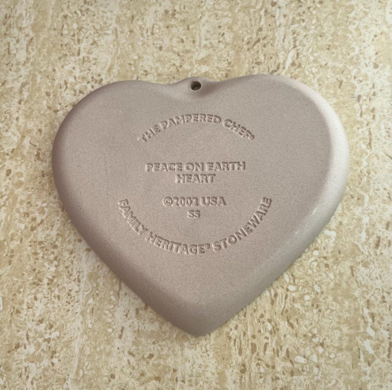 May include: A heart-shaped stoneware piece with the text "The Pampered Chef", "Peace on Earth Heart", and "Family Heritage Stoneware". The piece is a light beige colour and has a small hole at the top for hanging.