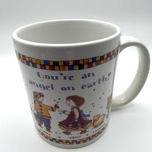 May include: A white ceramic mug featuring a colorful checkered border at the top and bottom. The text reads, You're an angel on earth. The illustration depicts two figures, one labeled Charity and one labeled Grace, with a small animal nearby.