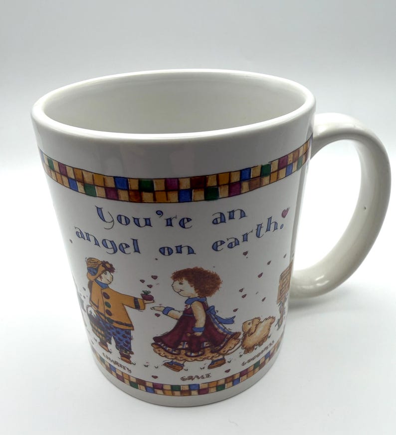 May include: A white ceramic mug featuring a colorful checkered border at the top and bottom. The text reads, You're an angel on earth. The illustration depicts two figures, one labeled Charity and one labeled Grace, with a small animal nearby.