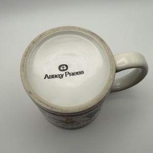 May include: A white ceramic mug viewed from above, featuring a handle on the right side. The bottom of the mug displays the black text Abbey Press with a small house icon above it. The mug has a smooth, glossy finish and a circular base.
