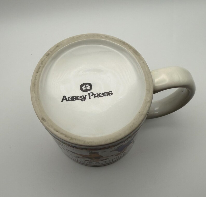 May include: A white ceramic mug viewed from above, featuring a handle on the right side. The bottom of the mug displays the black text Abbey Press with a small house icon above it. The mug has a smooth, glossy finish and a circular base.