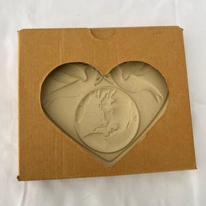 May include: A heart-shaped craft featuring a map of the British Isles and two doves, presented in a brown cardboard box with a heart-shaped window. The craft is a light beige colour.