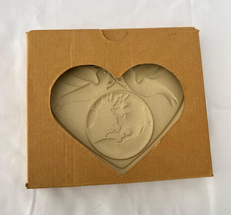 May include: A heart-shaped craft featuring a map of the British Isles and two doves, presented in a brown cardboard box with a heart-shaped window. The craft is a light beige colour.