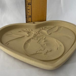 May include: Tan heart-shaped ceramic plaque with a relief design. The design features two doves, an olive branch, and a globe. The plaque is approximately 15 cm wide and has a small hole at the top for hanging.