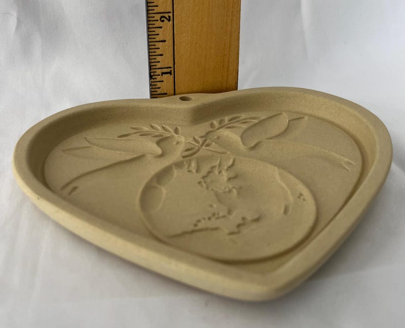 May include: Tan heart-shaped ceramic plaque with a relief design. The design features two doves, an olive branch, and a globe. The plaque is approximately 15 cm wide and has a small hole at the top for hanging.