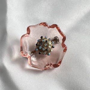 May include: A pink glass flower-shaped dish with a clear center, holding a silver pin with a round, multi-colored rhinestone design.