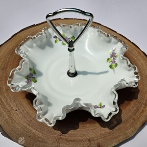 Puede incluir: Una fuente blanca con borde ondulado y asa plateada. La fuente presenta flores moradas y hojas verdes pintadas a mano. La fuente es probablemente de vidrio o porcelana y es un art&iacute;culo decorativo.