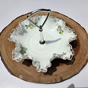 Puede incluir: Plato para servir blanco con borde ondulado y asa plateada. El plato presenta flores moradas y hojas verdes pintadas a mano. Se encuentra sobre una superficie redonda de madera, creando una presentaci&oacute;n r&uacute;stica y elegante. Ideal para servir postres o aperitivos.