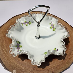 Puede incluir: Una fuente blanca con borde ondulado y asa plateada. El plato est&aacute; decorado con flores moradas y hojas verdes pintadas a mano. El plato probablemente sea de vidrio o porcelana y est&aacute; dise&ntilde;ado para servir alimentos o exhibir objetos decorativos.