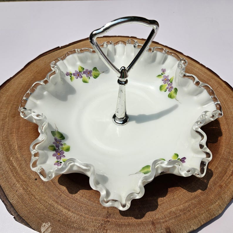 Puede incluir: Una fuente blanca con borde ondulado y asa plateada. El plato est&aacute; decorado con flores moradas y hojas verdes pintadas a mano. El plato probablemente sea de vidrio o porcelana y est&aacute; dise&ntilde;ado para servir alimentos o exhibir objetos decorativos.