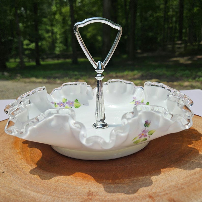 Puede incluir: Una fuente blanca de dos niveles con un asa plateada. El plato tiene un borde con volantes con un ribete plateado y est&aacute; decorado con detalles florales morados. El plato probablemente est&eacute; hecho de porcelana o cer&aacute;mica.