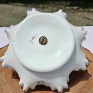 Puede incluir: Cuenco de vidrio blanco con borde ondulado y dise&ntilde;o floral. El cuenco tiene un borde festoneado con ribete plateado. La base del cuenco tiene el texto "J Brown" y un logotipo. El cuenco est&aacute; sobre una superficie de madera.