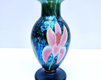 Puede incluir: Un jarrón decorativo de vidrio con base y cuerpo azul oscuro, con un diseño de flor rosa. El jarrón tiene un patrón azul moteado y un borde verde. La flor tiene pétalos rosas y un centro rojo.