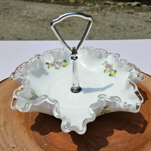 Puede incluir: Una fuente blanca con borde ondulado y asa plateada. La fuente presenta detalles florales p&uacute;rpuras y verdes pintados a mano. El asa es de metal plateado. La fuente est&aacute; sobre una superficie de madera.