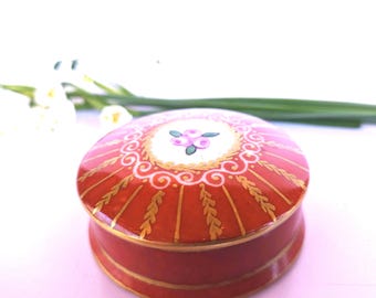 Puede incluir: Una pequeña caja de baratijas redonda y roja con un borde dorado y un diseño floral en la tapa. La caja está decorada con un patrón blanco y dorado.