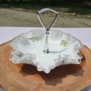 Puede incluir: Plato para servir blanco con borde con volantes y asa plateada. El plato presenta detalles florales morados pintados a mano. El asa es de metal plateado. El plato est&aacute; sobre una superficie de madera.