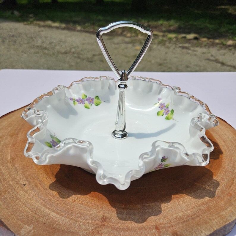 Puede incluir: Plato para servir blanco con borde con volantes y asa plateada. El plato presenta detalles florales morados pintados a mano. El asa es de metal plateado. El plato est&aacute; sobre una superficie de madera.