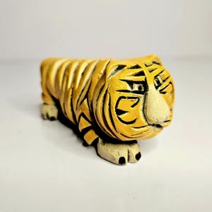 Peut inclure: Une figurine de tigre en céramique avec un motif rayé jaune et brun. Le tigre a une expression de dessin animé avec un gros nez et de petits yeux.