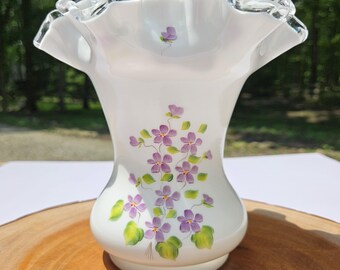 Puede incluir: Jarrón de cerámica blanca con borde con volantes y diseño floral morado pintado a mano. El jarrón tiene una parte superior ancha y acampanada y una base redondeada. Las flores están acentuadas con hojas verdes. Una pieza decorativa para la decoración del hogar.