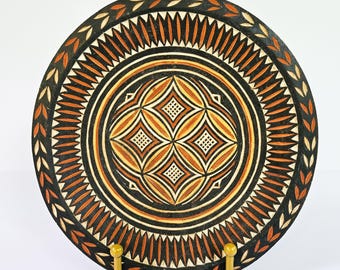 Puede incluir: Un plato decorativo redondo de madera con un diseño intrincado. El plato presenta un fondo negro con patrones geométricos de color naranja y crema. El borde exterior tiene un patrón en forma de hoja. El plato se exhibe sobre un soporte amarillo.