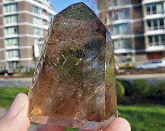 May include: A faceted, smoky quartz crystal point. The crystal is a translucent brown color with visible internal structures. The crystal is held up against a bright blue sky and buildings in the background.