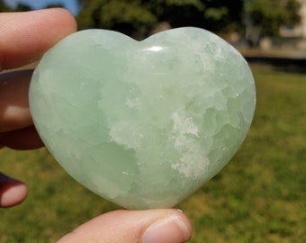 May include: A pale green heart-shaped crystal with a smooth, polished finish. The stone has a slightly translucent quality and a textured, crystalline pattern. The heart is held in a hand against a background of green grass, trees, and a blue sky.